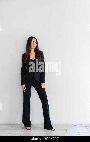 Femme regardant côté en pantalon ou costume formel noir se tient contre le mur blanc dans le studio. Modèle brune caucasien avec les cheveux longs. Portrait professionnel Banque D'Images
