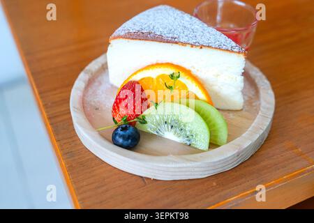 Une tranche de cheesecake crémeux nappée de sucre en poudre et accompagnée de fruits frais incluant fraise, myrtille, kiwi et orange sur un WO rond Banque D'Images