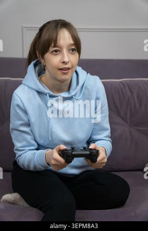 Gamer Girl assis confortablement sur un canapé confortable, tenant une manette et se plongeant dans des jeux vidéo passionnants, profitant du temps de loisirs à la maison et e Banque D'Images
