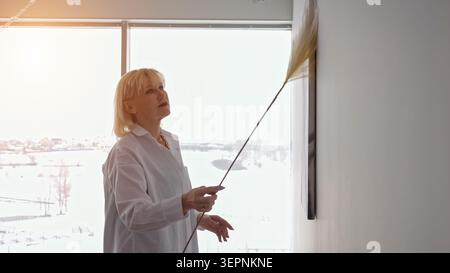 Femme senior en chemise blanche dépoussiérant des œuvres d'art murales avec un plumeau dans un salon moderne lumineux, axé sur le maintien de sa maison bien rangée et Banque D'Images