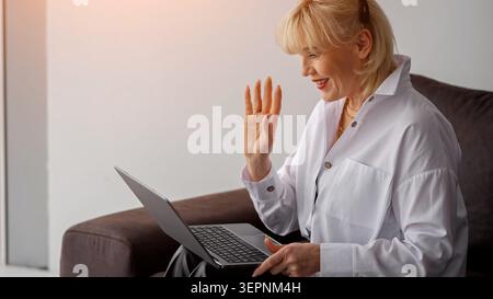 Femme âgée heureuse assise sur un canapé à la maison, souriant et agitant pendant un appel vidéo d'ordinateur portable pour rester connecté avec la famille et les amis, détendu et Banque D'Images