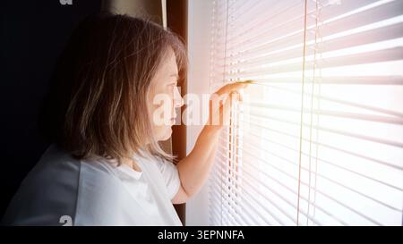 Femme âgée debout près d'une fenêtre, regardant à travers des stores horizontaux avec ses doigts, réfléchissant sur les concepts de solitude, d'isolement et d'espoir Banque D'Images