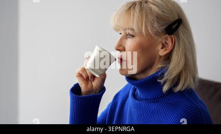 Femme mature dans un col roulé bleu sirotant d'une tasse blanche, trouvant la paix et la détente au cours de sa routine tôt le matin, créant un concept de Banque D'Images
