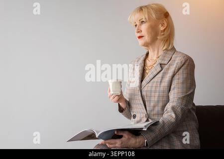 Femme blonde mature portant un blazer à carreaux, tenant un magazine et une tasse, regardant loin contemplativement, perdu dans la pensée en attendant ou en prenant un Banque D'Images
