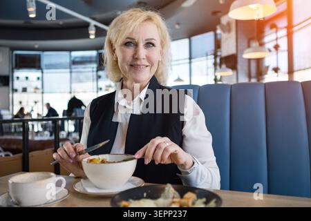 Senior femme professionnel savourant un repas équilibré l'après-midi avec ordinateur portable à proximité. Femme âgée en tenue d'affaires souriante tout en mangeant un déjeuner nutritif Banque D'Images