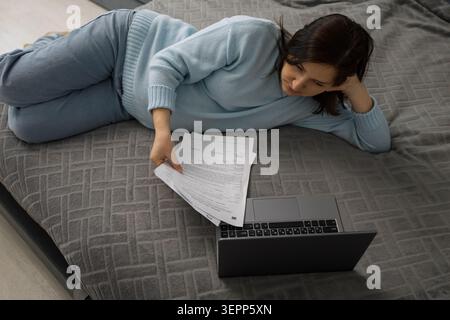 Femme enceinte allongée sur un lit dans une chambre confortable tout en regardant des documents à côté d'un ordinateur portable ouvert, scène de maison détendue avec une lumière naturelle douce et grise Banque D'Images