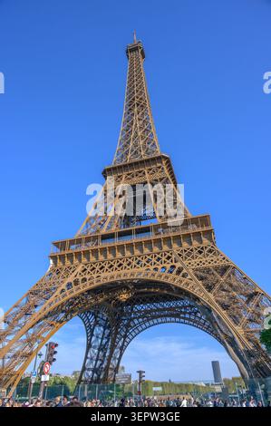 Paris, France - 04 avril 2026 : vue en bas angle de la Tour Eiffel avec foule et panneaux de signalisation sous un ciel bleu clair. Banque D'Images