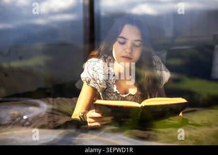 Jeune femme lisant le livre dans la lumière chaude de la fenêtre. Jeune femme est allongée sur un lit lisant un livre ouvert à côté d'une grande fenêtre, avec une lumière chaude du soleil, douce Banque D'Images