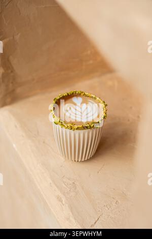 Un latte chaud à la pistache présentant un élégant latte à feuilles, servi dans une tasse blanche nervurée avec un bord de pistache écrasé sur une surface texturée. Banque D'Images