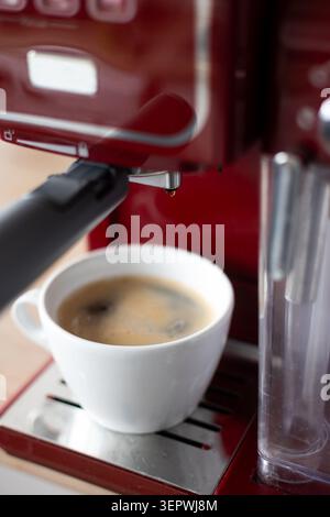 Gros plan d'un espresso fraîchement préparé servi dans une tasse en céramique blanche sous une machine à espresso rouge. Banque D'Images