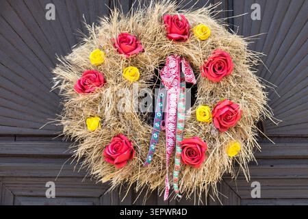 Couronne de paille traditionnelle du festival des récoltes de Moravie du Sud décorée de fleurs artificielles de Rose Rosa et de rubans folkloriques sur une porte en bois sombre Banque D'Images