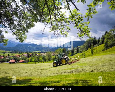 Tracteur vert fauchant prairie alpine avec les montagnes des Alpes Allgau en arrière-plan - paysage rural d'été, Bavière, Allemagne Banque D'Images