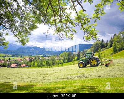 Tracteur vert fauchant prairie alpine avec les montagnes des Alpes Allgau en arrière-plan - paysage rural d'été, Bavière, Allemagne Banque D'Images