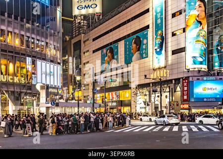 Des foules attendent au passage piéton Shibuya Scramble sous les immeubles commerciaux et les panneaux publicitaires illuminés la nuit à Tokyo, au Japon. Banque D'Images