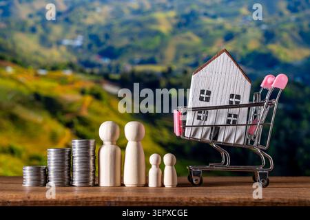 Figurines de famille en bois et pièces empilées symbolisant l'épargne et la croissance financière, avec une petite maison dans un panier représentant l'achat d'une maison et Banque D'Images