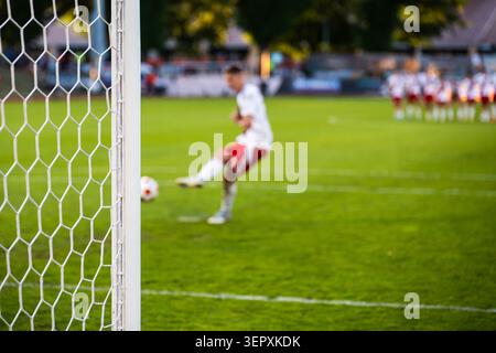 Un joueur de football dans un uniforme blanc frappant un tir de pénalité vers le but pendant un match de football, vu à travers le filet. Banque D'Images