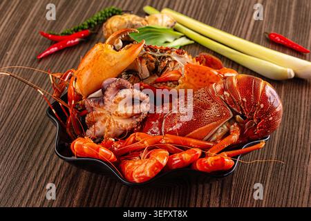 Plateau de fruits de mer frais avec du crabe de poulpe de homard et des crevettes servi dans un bol noir sur une table en bois avec des piments et de la citronnelle pour la cuisson Banque D'Images