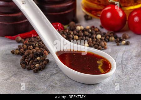 Cuillère en céramique blanche remplie de sauce épicée au Chili rouge, entourée de grains de poivre, de piments rouges séchés et de tomates fraîches sur une surface grise texturée Banque D'Images