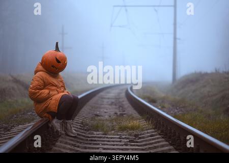 Personnage mystérieux avec un jack o tête de lanterne assis sur les voies ferrées dans la brume épaisse. Scène thématique d'Halloween alliant solitude, mystère et narration cinématographique Banque D'Images
