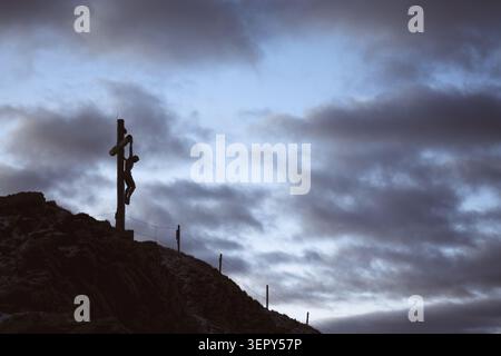 Crucifix de Jésus-Christ silhouetté contre un ciel nuageux dramatique dans les Dolomites. Paysage montagneux atmosphérique au crépuscule avec des paysages alpins accidentés Banque D'Images