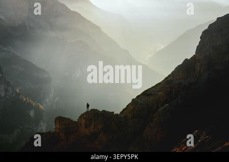 Un randonneur solitaire se tient sur une crête rocheuse au-dessus de Cortina d'Ampezzo au lever du soleil dans les Dolomites. Paysage montagneux spectaculaire avec rayons de soleil, brume et sommets alpins en couches Banque D'Images