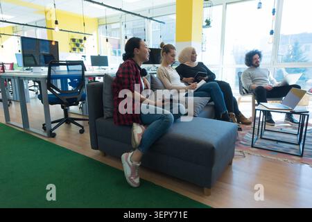 Plan d'ensemble d'une équipe détendue mais productive de jeunes professionnels divers assis sur un canapé et des chaises, utilisant des ordinateurs portables et des tablettes dans un Banque D'Images