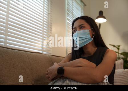 Femme asiatique portant un masque facial protecteur assis sur le canapé à la maison pour la quarantaine Banque D'Images