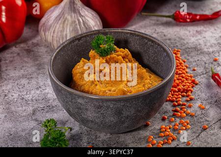 Purée d'orange épaisse à base de lentilles rouges servie dans un bol gris rustique, entourée de lentilles sèches éparpillées, de bulbe d'ail frais, de poivrons rouges et de chi Banque D'Images