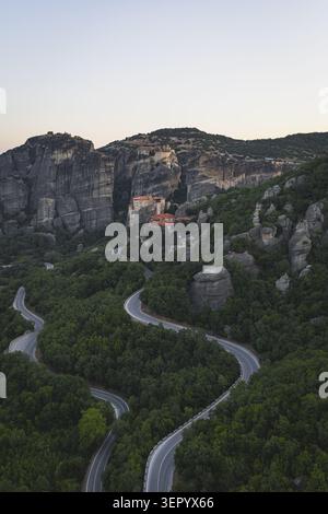Vue aérienne des monastères des Météores perchés sur d'imposants piliers de roche avec une route de montagne sinueuse à travers des forêts vertes denses au crépuscule à Trikala, G. Banque D'Images