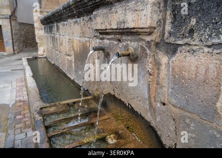 Vue rapprochée d'une fontaine publique en pierre altérée avec deux becs métalliques libérant un jet d'eau régulier dans un creux en dessous, situé dans le village Banque D'Images