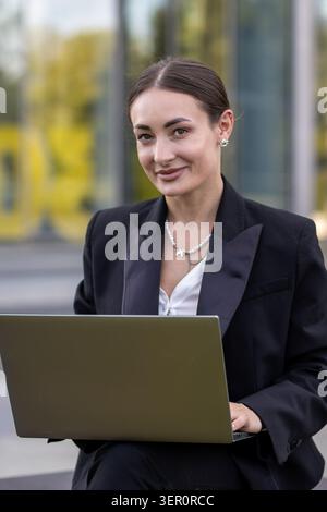 Une femme aux cheveux longs est assise dehors avec un ordinateur portable sur ses genoux. Elle est habillée de vêtements formels et sourit en regardant l'écran. L'arrière-plan Banque D'Images
