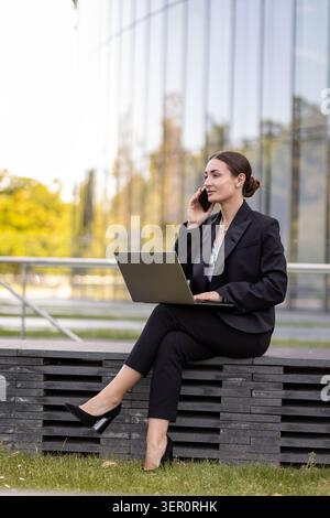Une femme est assise sur une pile de pierres sombres faisant un appel téléphonique. Elle utilise un ordinateur portable sur ses genoux. Elle est dehors près d'un grand bâtiment de verre par une journée ensoleillée. Banque D'Images
