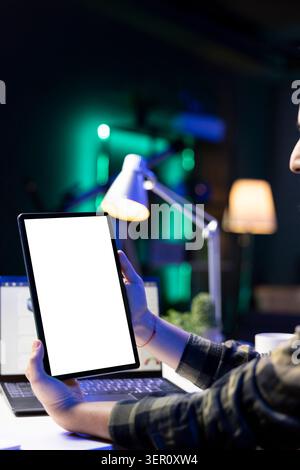 Personne tenant verticalement la tablette avec écran blanc devant l'ordinateur portable. Freelance masculine travaillant à la maison, axée sur l'appareil numérique affichant un modèle vierge de maquette de clé chroma. Banque D'Images