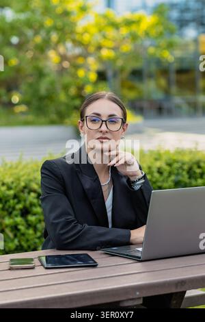 Une femme s'assoit à une table à l'extérieur en utilisant un ordinateur portable. Elle est vêtue d'une tenue d'affaires et semble s'engager dans son travail. Le cadre a de la verdure et natura Banque D'Images