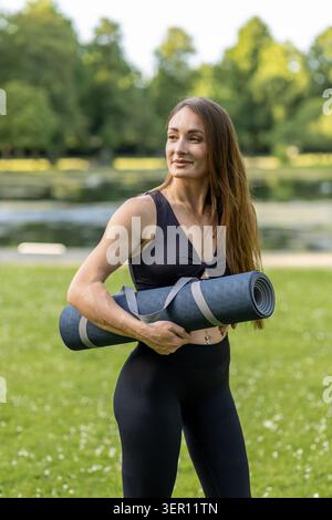 Une femme se tient debout dans un parc tenant un tapis de yoga. Elle a les cheveux longs et porte une tenue noire. De l'herbe verte l'entoure et il y a de l'eau dans le backgroun Banque D'Images