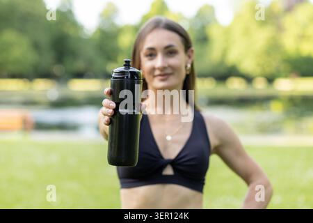 Une jeune femme se tient dans un parc par une journée ensoleillée. Elle pose avec une bouteille d'eau noire dans une main. Les arbres verts et l'herbe l'entourent comme elle sourit a Banque D'Images