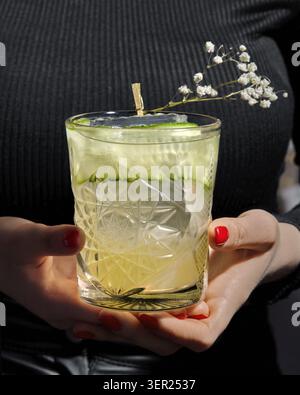 Femme barman servant un verre de cristal de gin rafraîchissant et cocktail tonique décoré de tranches de concombre et de branche de gypsophila Banque D'Images