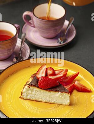 Tranche de cheesecake au chocolat et aux fraises sur une assiette jaune, avec deux tasses de thé sur une table noire et le thé versé Banque D'Images