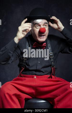Portrait d'un clown sur fond sombre. Un homme âgé avec un nez de clown rouge et un chapeau melon sur la tête. Vêtu d'un pantalon rouge et d'une chemise noire avec suspension Banque D'Images
