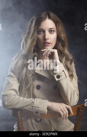 Portrait de la jeune belle femme blonde est assise sur une chaise. Femelle en manteau beige sur fond de fumée Banque D'Images