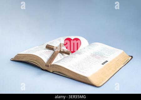 Croix de Crucifx et coeur de papier rouge sur le dessus de la Sainte Bible. Isolé sur fond bleu clair. Banque D'Images