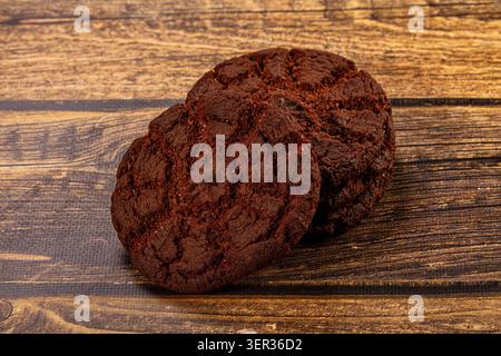 Deux biscuits au chocolat noir reposant sur une surface en bois rustique avec des tons bruns riches et une texture froissée. Un éclairage chaleureux met en valeur les détails des produits de boulangerie. Banque D'Images