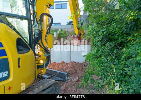 Une mini-pelle jaune fonctionne dans une cour arrière pendant les travaux d'aménagement paysager, installant des blocs de mur de soutènement en béton près d'une colline verdoyante. Compact Banque D'Images