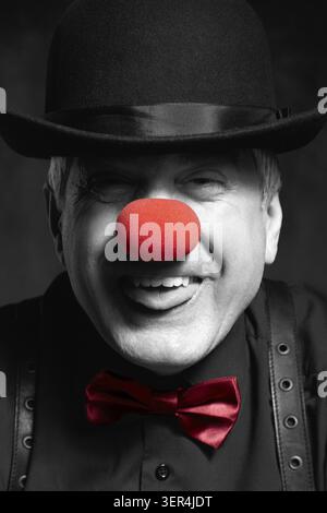 Portrait couleur sélective d'un clown avec un chapeau tiré vers le bas sur ses yeux sort de sa langue. Un homme âgé avec un nez de clown rouge et un chapeau melon sur h. Banque D'Images