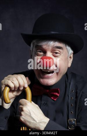 Portrait de clown riant sur un fond sombre appuyé sur le manche d'un parapluie. Un homme âgé avec un nez de clown rouge et un chapeau melon sur la tête. Banque D'Images