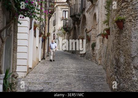 Homme âgé marchant le long d'une rue pavée étroite bordée de maisons en pierre et de fleurs dans le centre historique de Gerace, parc national d'Aspromonte Banque D'Images