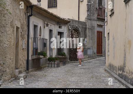 Femme en robe rose et chapeau blanc marchant le long d'une rue pavée étroite dans le centre historique de Gerace, Aspromonte National Park, Calabre Banque D'Images