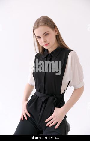 Une jeune femme blonde avec ses mains sur ses hanches dans une combinaison noire sans manches sur une blouse blanche à manches bouffantes dans un studio Banque D'Images
