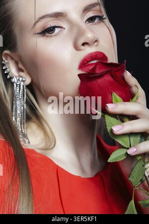 Femelle avec rose rouge à la main et longue boucle d'oreille avec strass dans l'oreille. Portrait d'une jeune femme en robe rouge et un brin de cheveux sur le visage Banque D'Images
