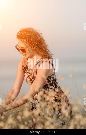 Femme champ d'été belle femme rousse portant des lunettes de soleil debout dans la robe florale parmi les fleurs sauvages profitant de l'atmosphère de coucher de soleil pendant chaud Banque D'Images